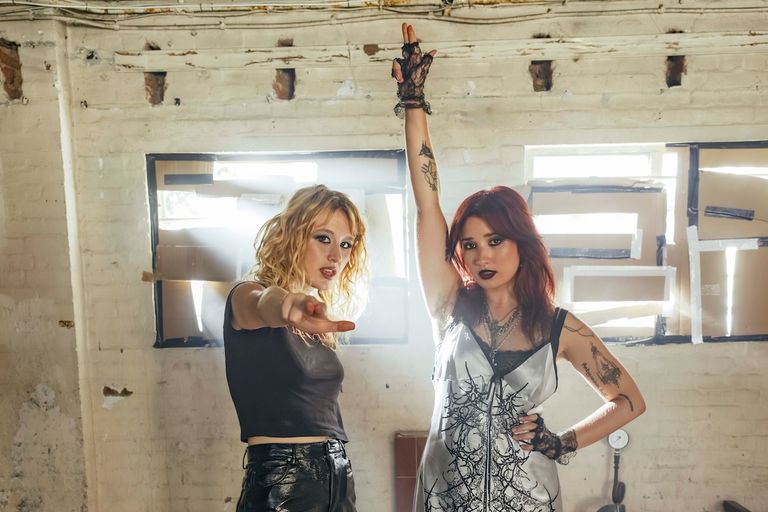 Punk duo Lambrini Girls posing in an industrial space with one member pointing forward and the other raising a gloved hand for a press photo.